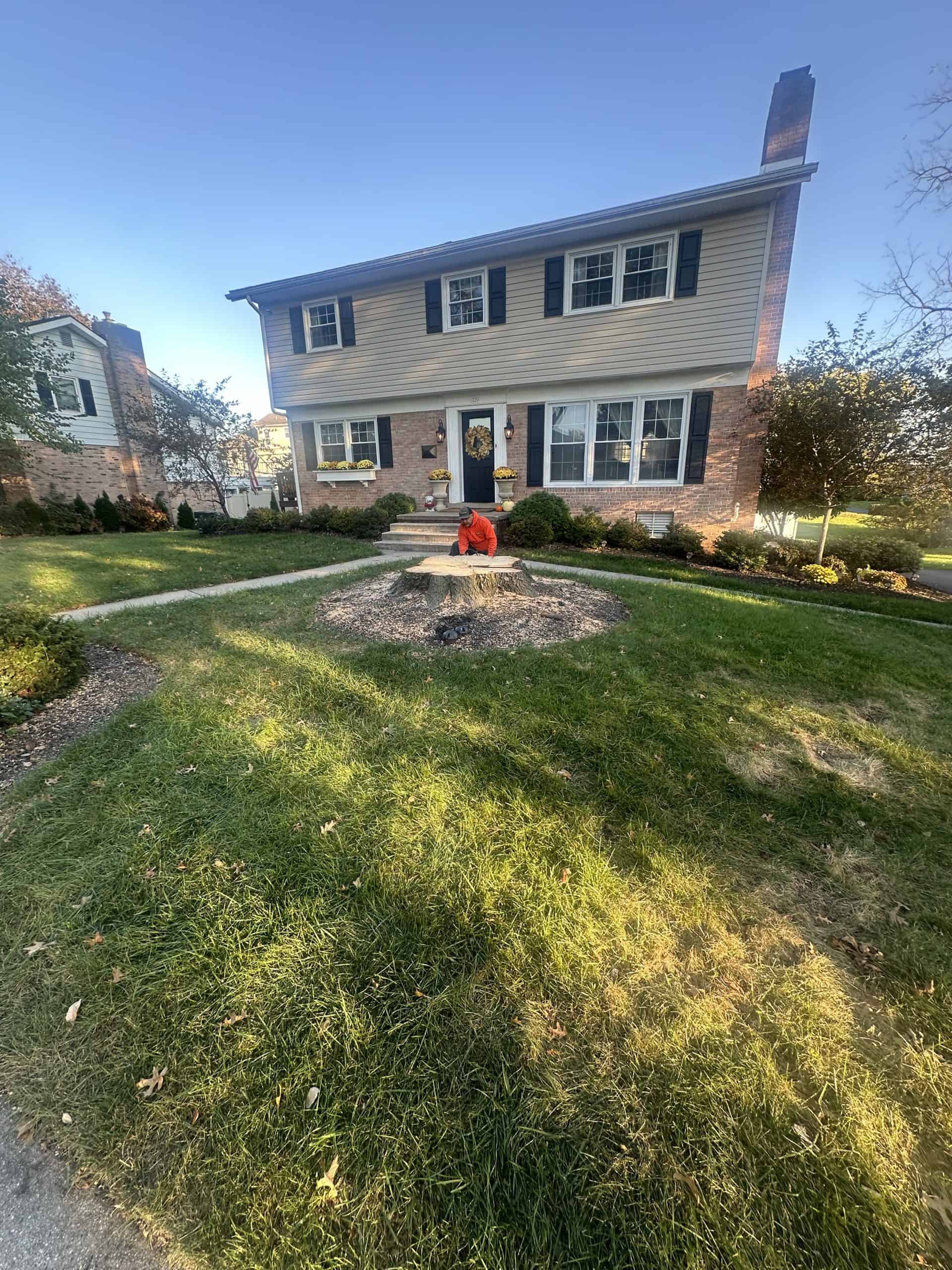 A child in a red jacket sits on a tree stump in the front yard of a two-story house with beige siding and brick accents. The yard is grassy with decorative shrubs near the house. The sky is clear and blue.